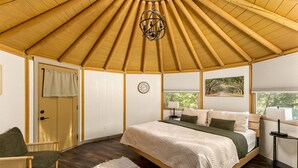 Un dormitorio con techo de madera, una cama con colcha blanca, una mesita de noche con lámpara, una ventana con vistas a árboles y una puerta con cortina.
