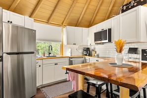 A modern kitchen with white cabinets, stainless steel appliances, a wooden island, and a view of greenery outside.