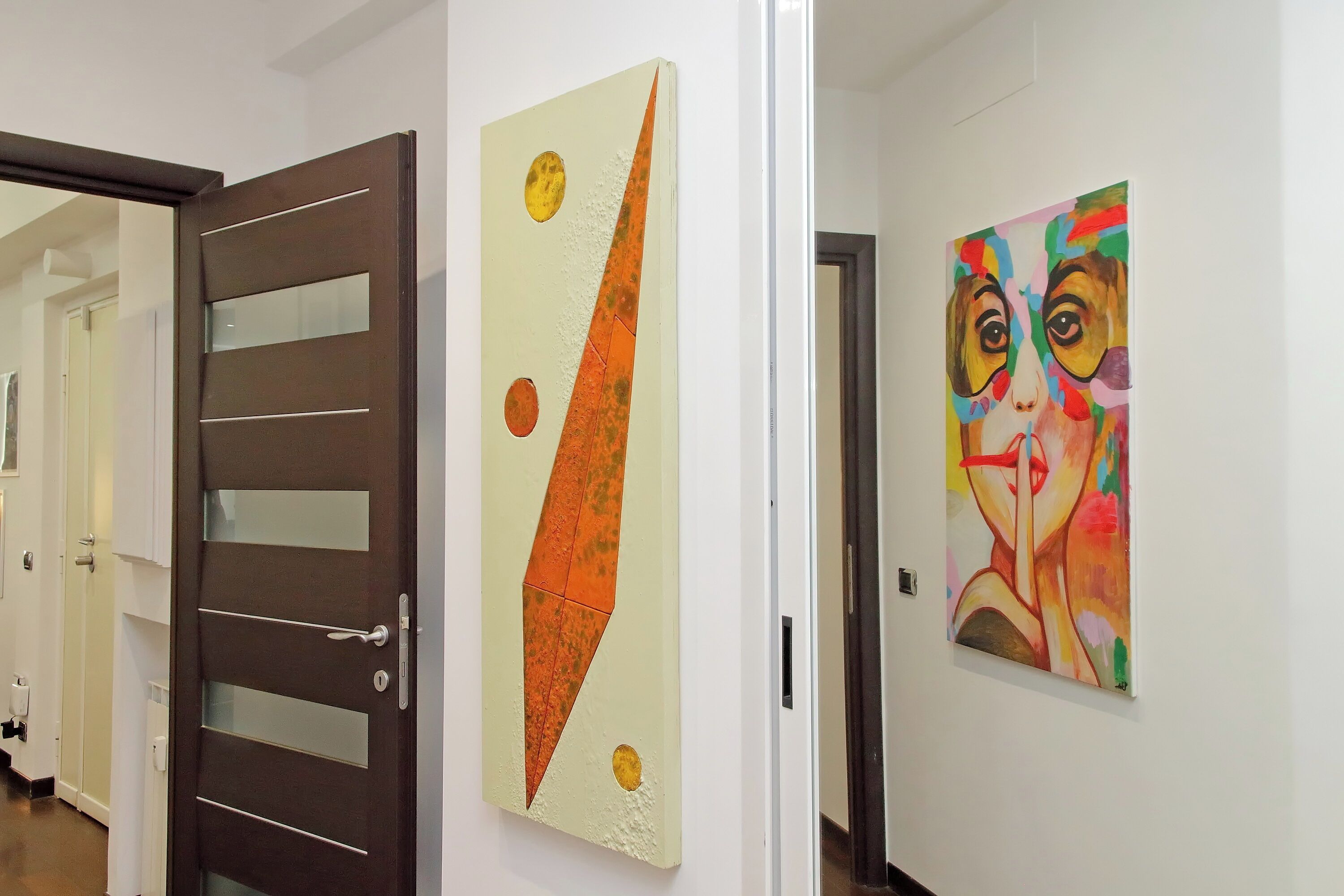 Un pasillo con una puerta moderna, un cuadro de formas geométricas y otro cuadro con figuras humanas abstractas.