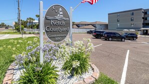 Une enseigne indiquant l’adresse de l’ensemble résidentiel « Ketch Courtyard Condominiums », avec un drapeau, entourée de fleurs et de voitures garées.