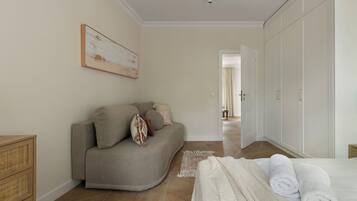 A bedroom with a sofa, a bed with rolled towels, a wicker chest of drawers, and a large wardrobe.