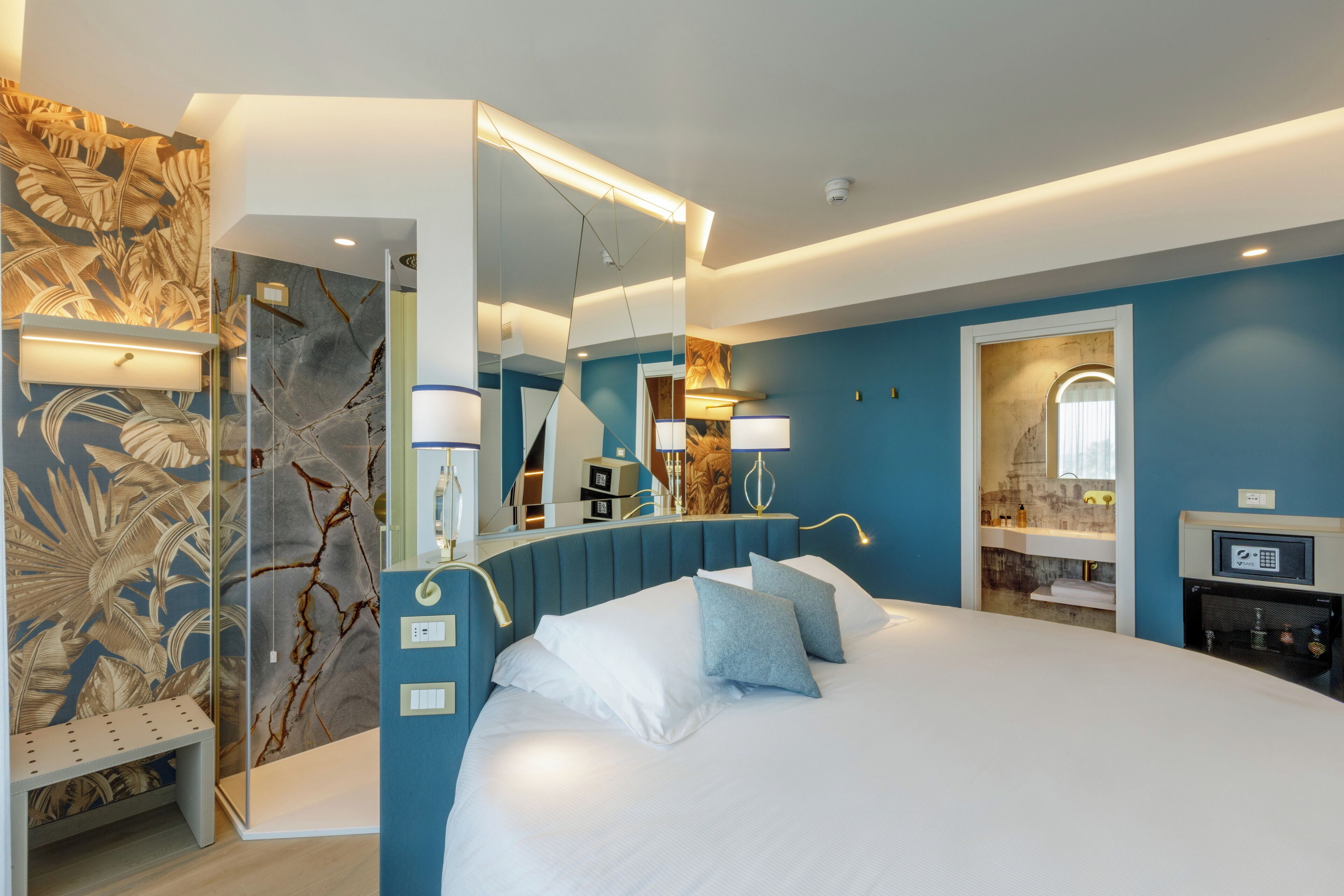 A modern hotel room with a large bed, a mirror, a vanity with a sink, and a bench.