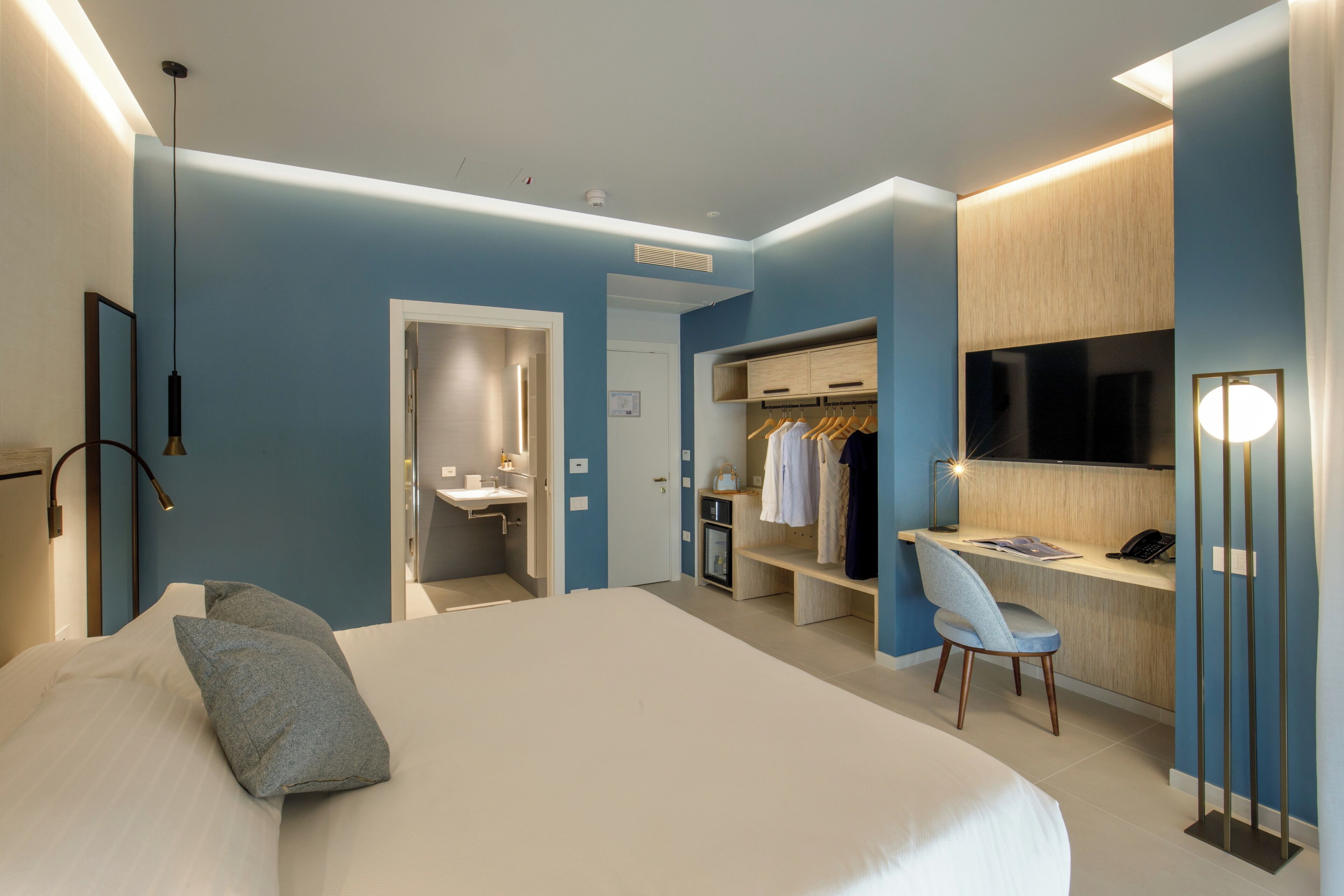 A modern hotel room with a large bed, a desk with a chair, a wardrobe, and a TV.