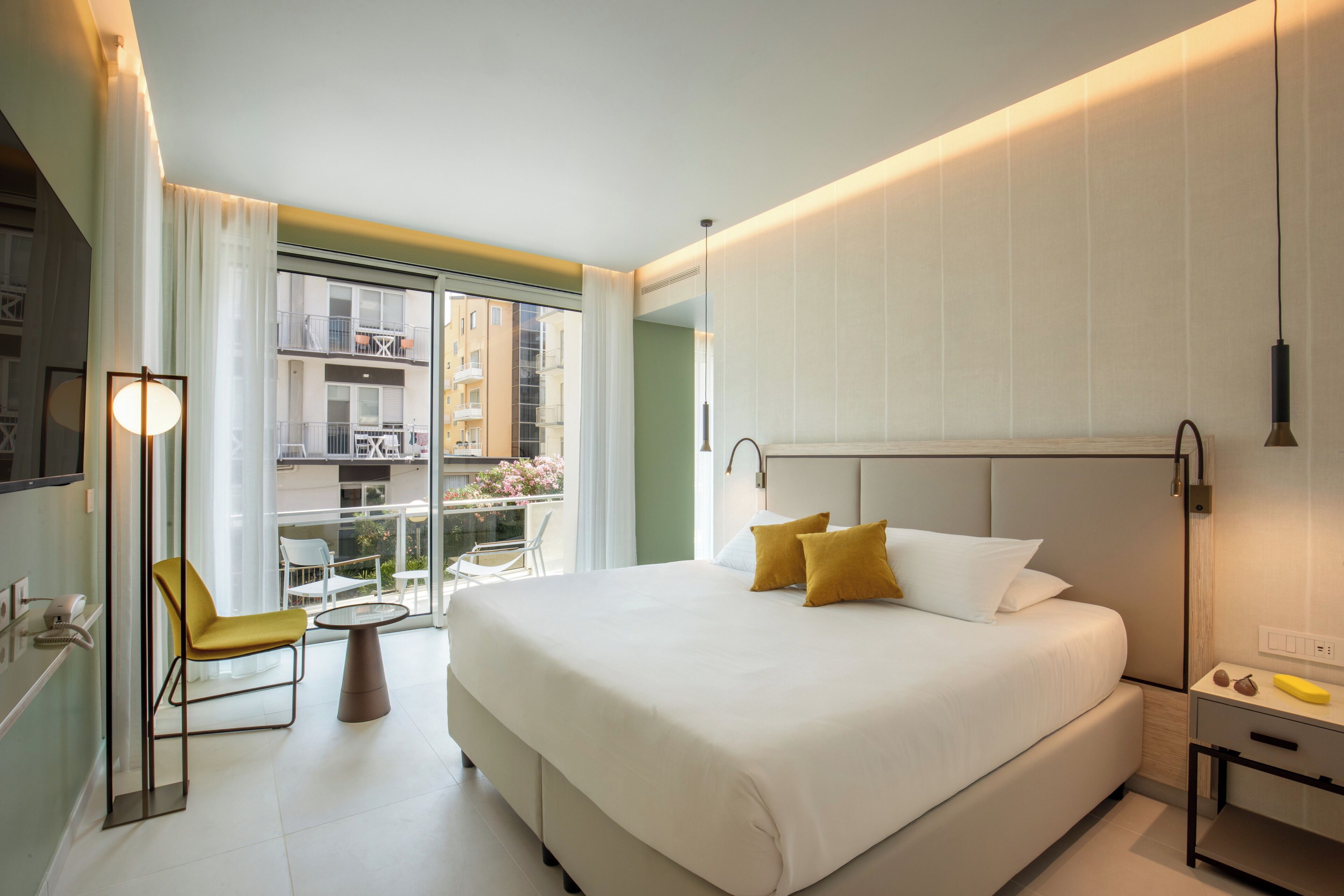 A modern hotel room with a large bed, a small table, a chair, and a balcony with a view of other buildings.