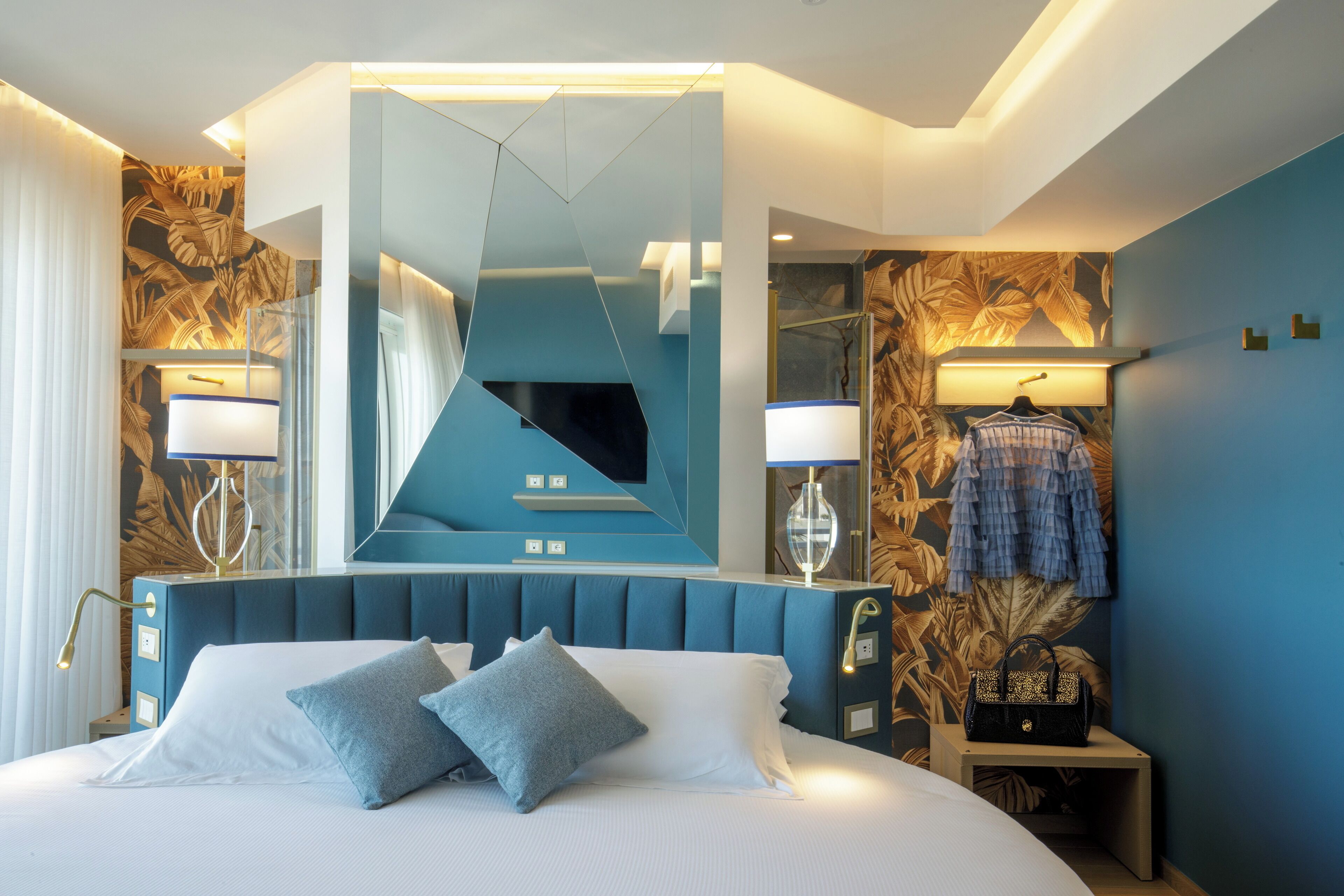 A modern hotel room with a large bed, a mirror, a nightstand, and a wall with a tropical leaf pattern.