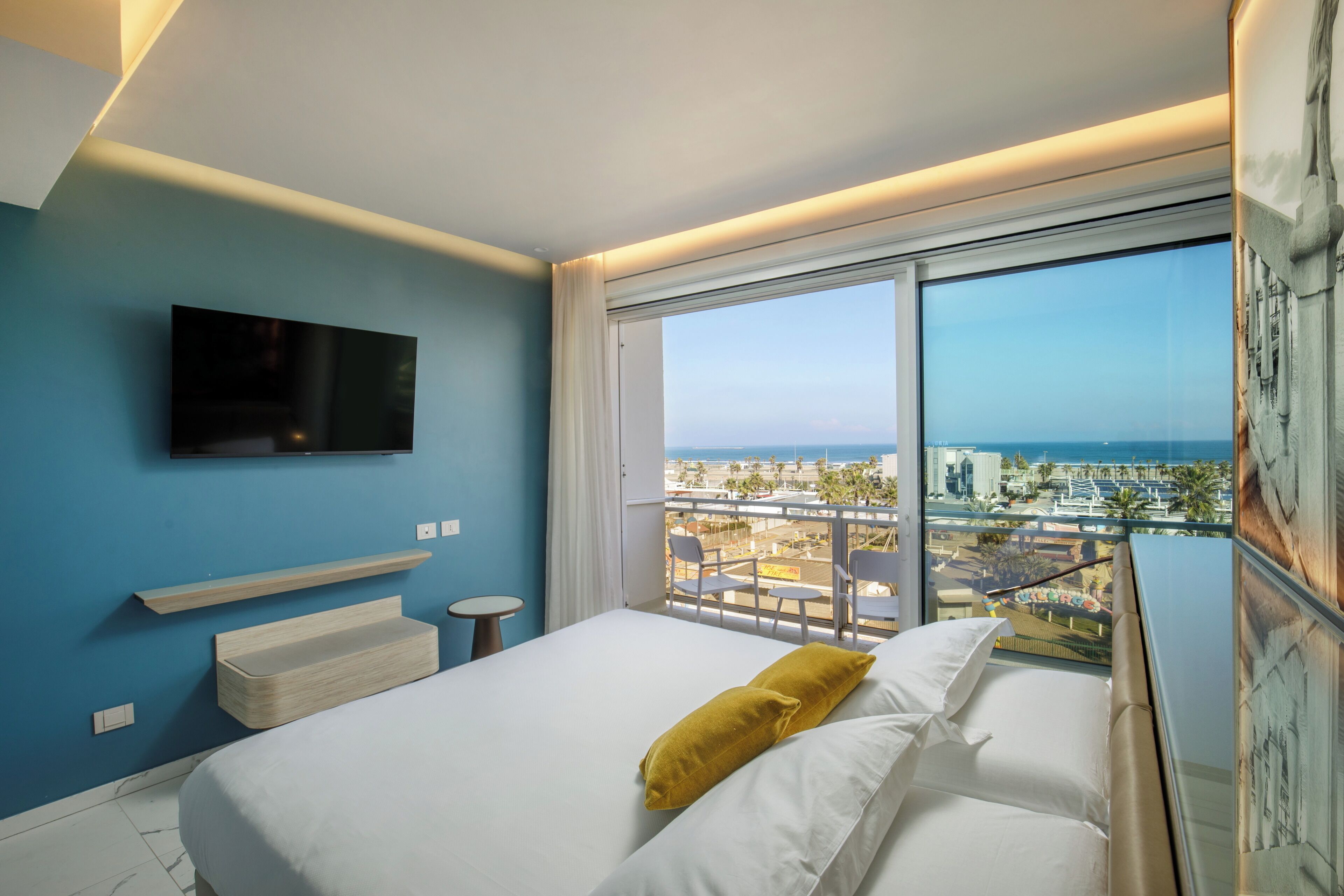 Habitación de hotel con cama, televisor montado, balcón con vistas a la ciudad y al mar, y un ventanal grande.