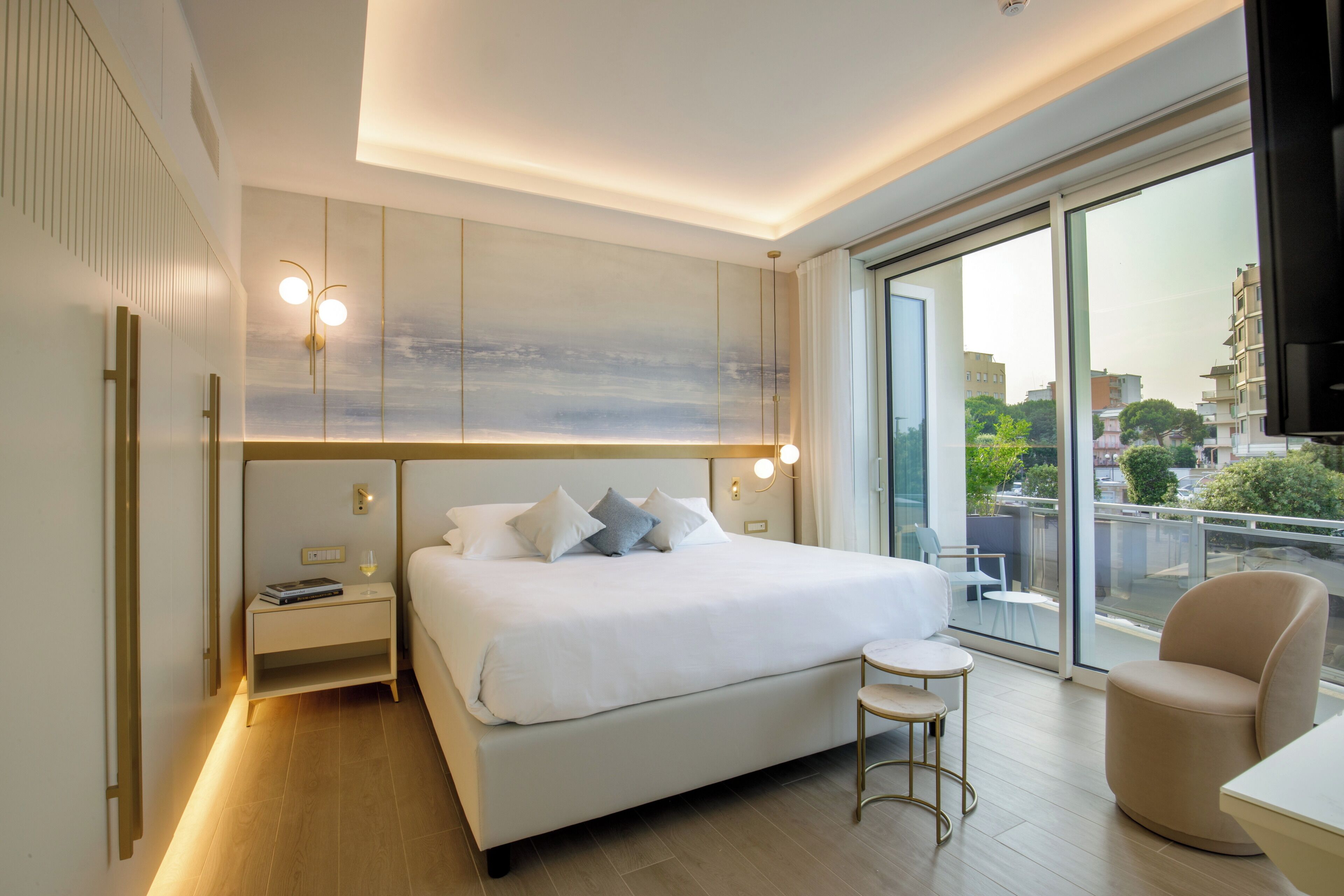 A modern hotel room with a large bed, a small round table, a chair, and a large window offering a city view.
