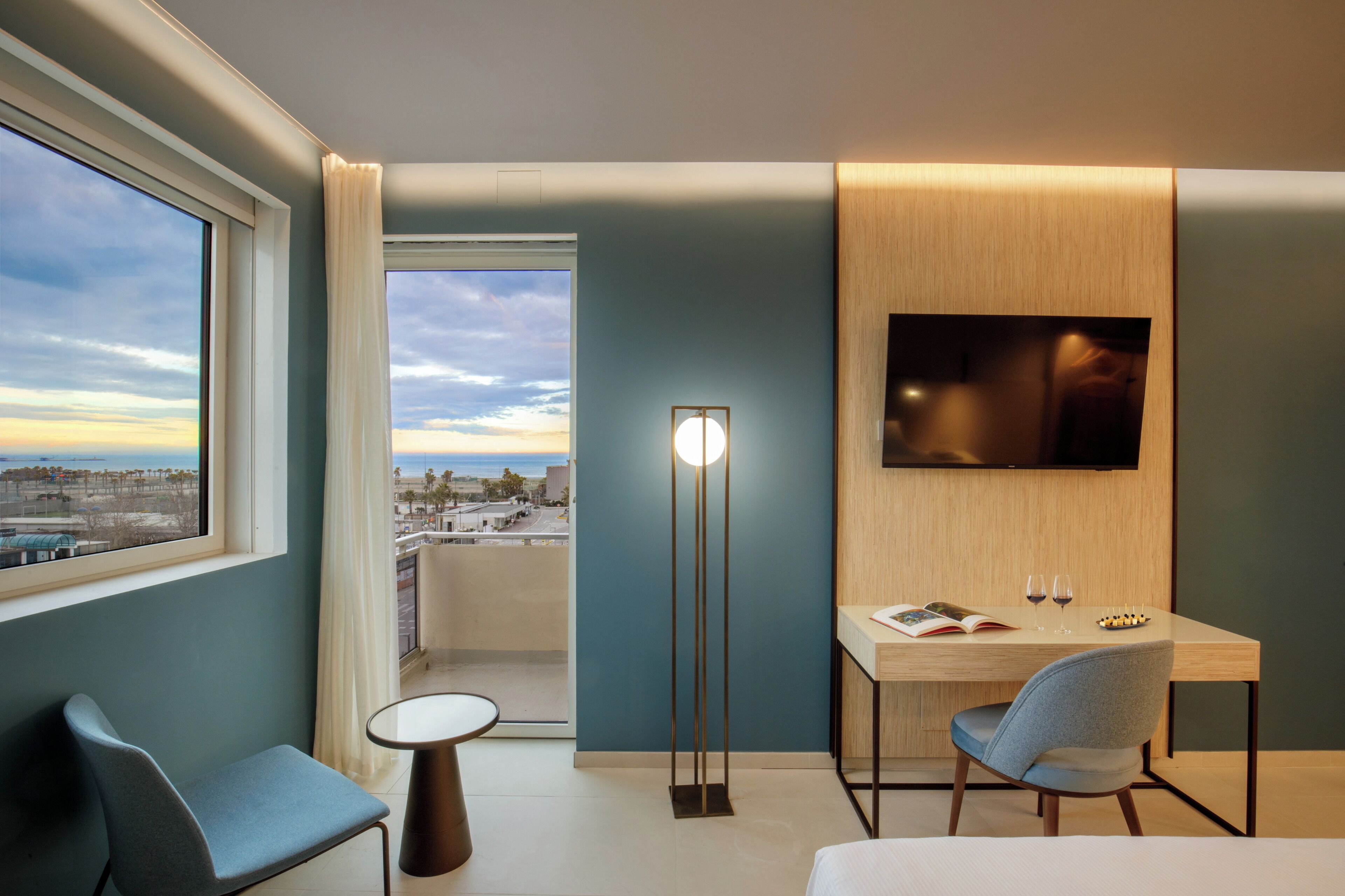 A modern hotel room with a balcony, a flat-screen TV, a desk with a chair, and a small table.