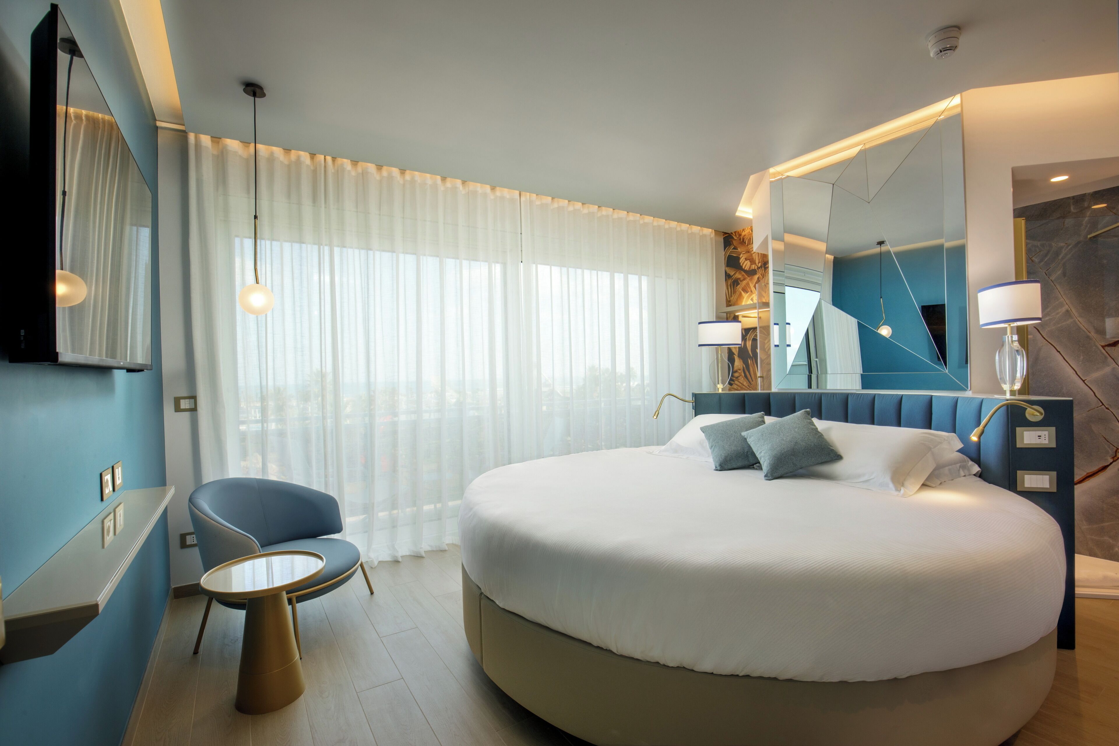 Una habitación de hotel moderna con una cama redonda, una mesita pequeña y redonda, un sillón azul y un espejo grande.