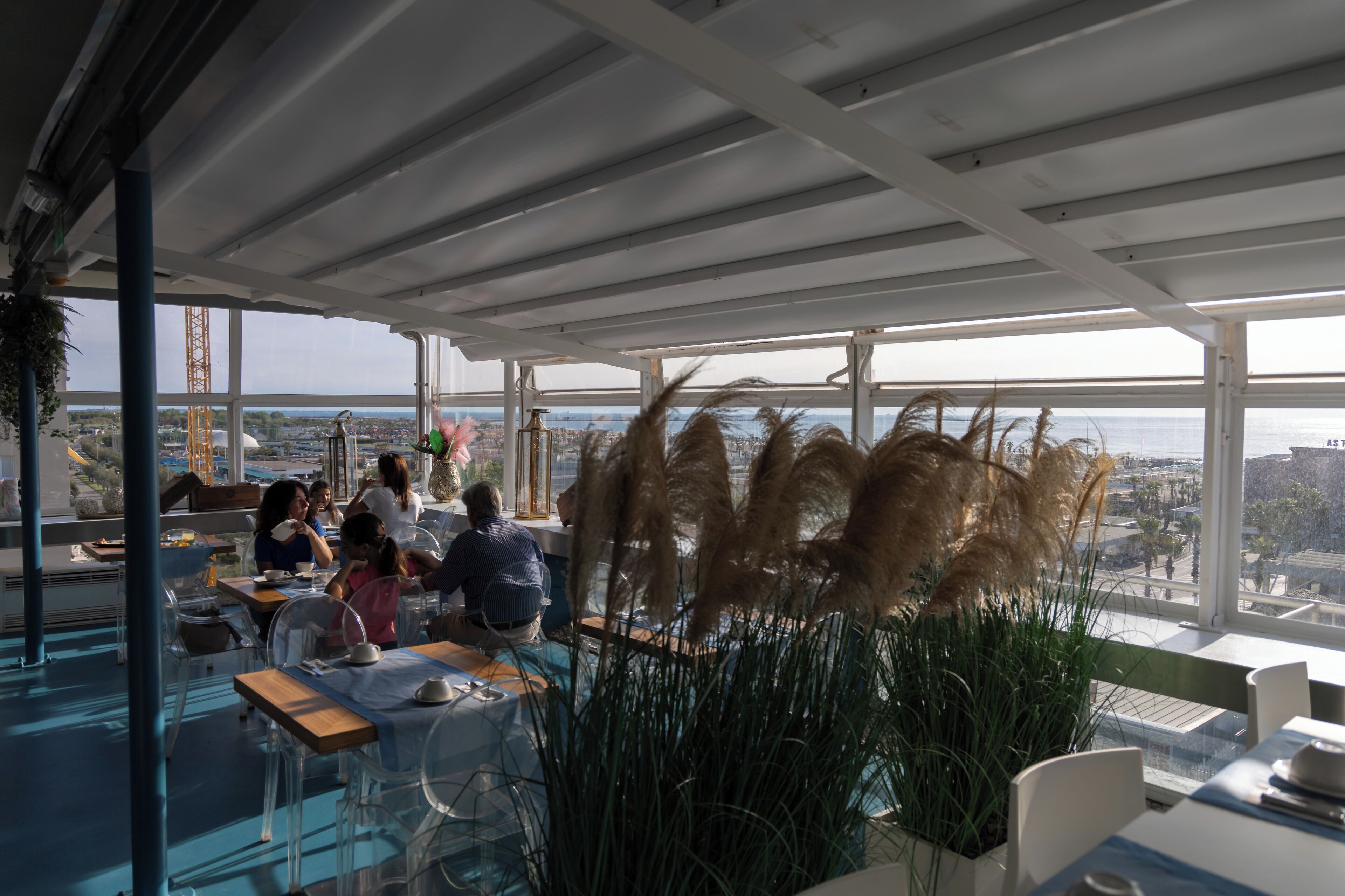 A modern restaurant with large windows offering a city view, clear glass tables, and potted plants.