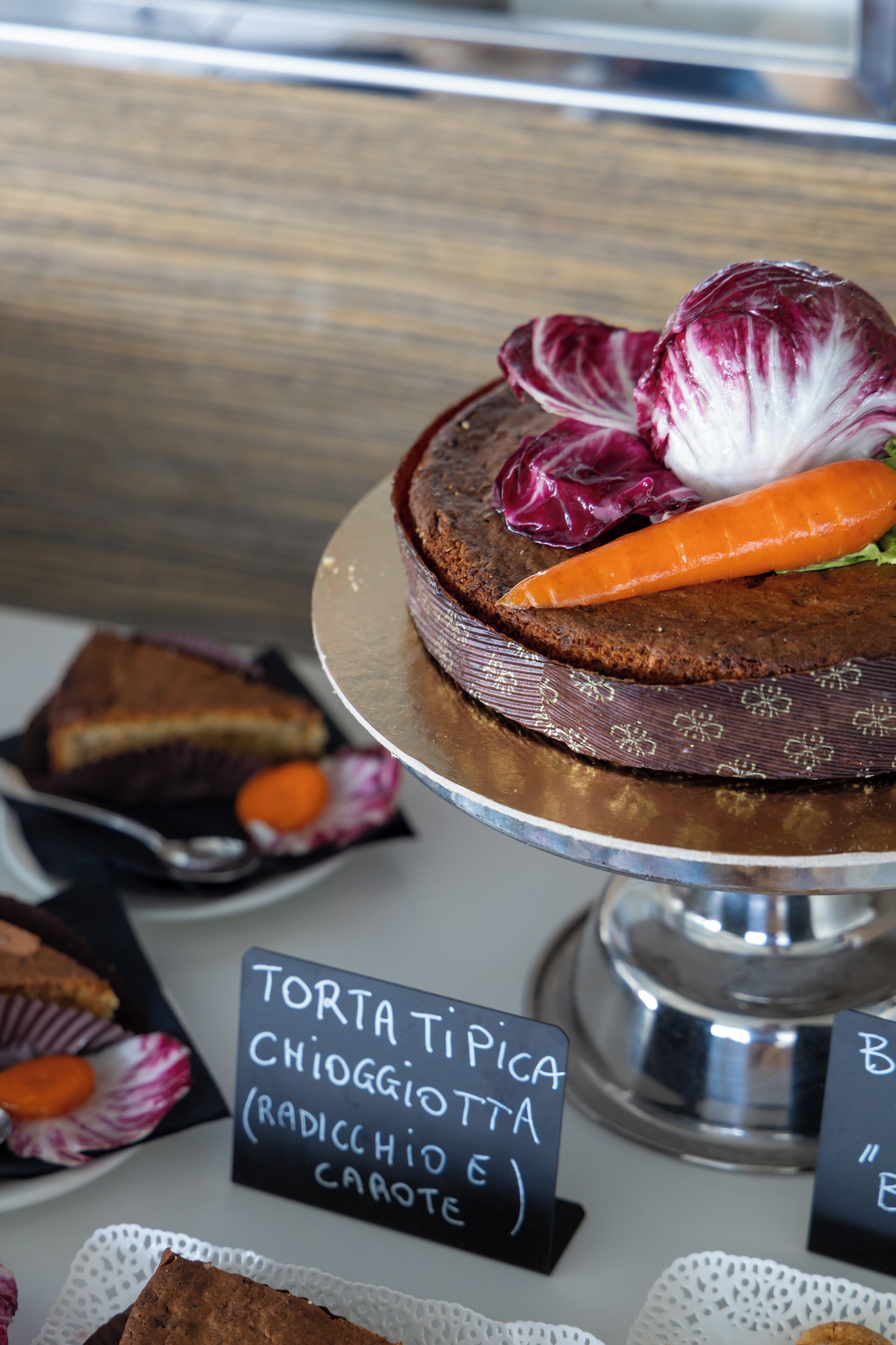 Une assiette de dessert avec un gâteau, une carotte et une feuille de radicchio, portant l’étiquette « TORTA TIPA CHIOGGIOTTA (RADICCHIO E CAROTE) ».