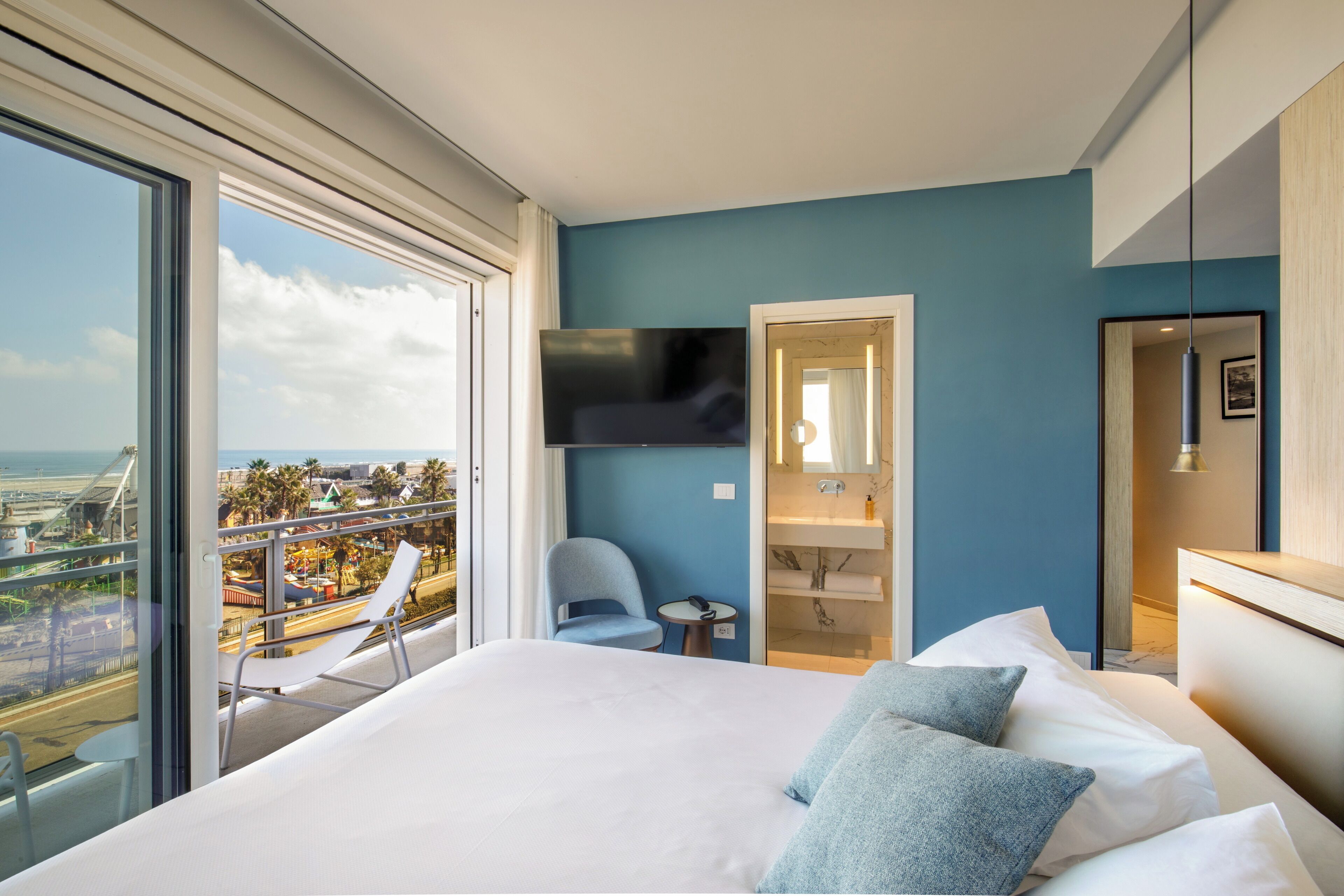 Una habitación de hotel moderna con una cama grande, un televisor montado en la pared, un balcón con una silla y vistas a la ciudad.
