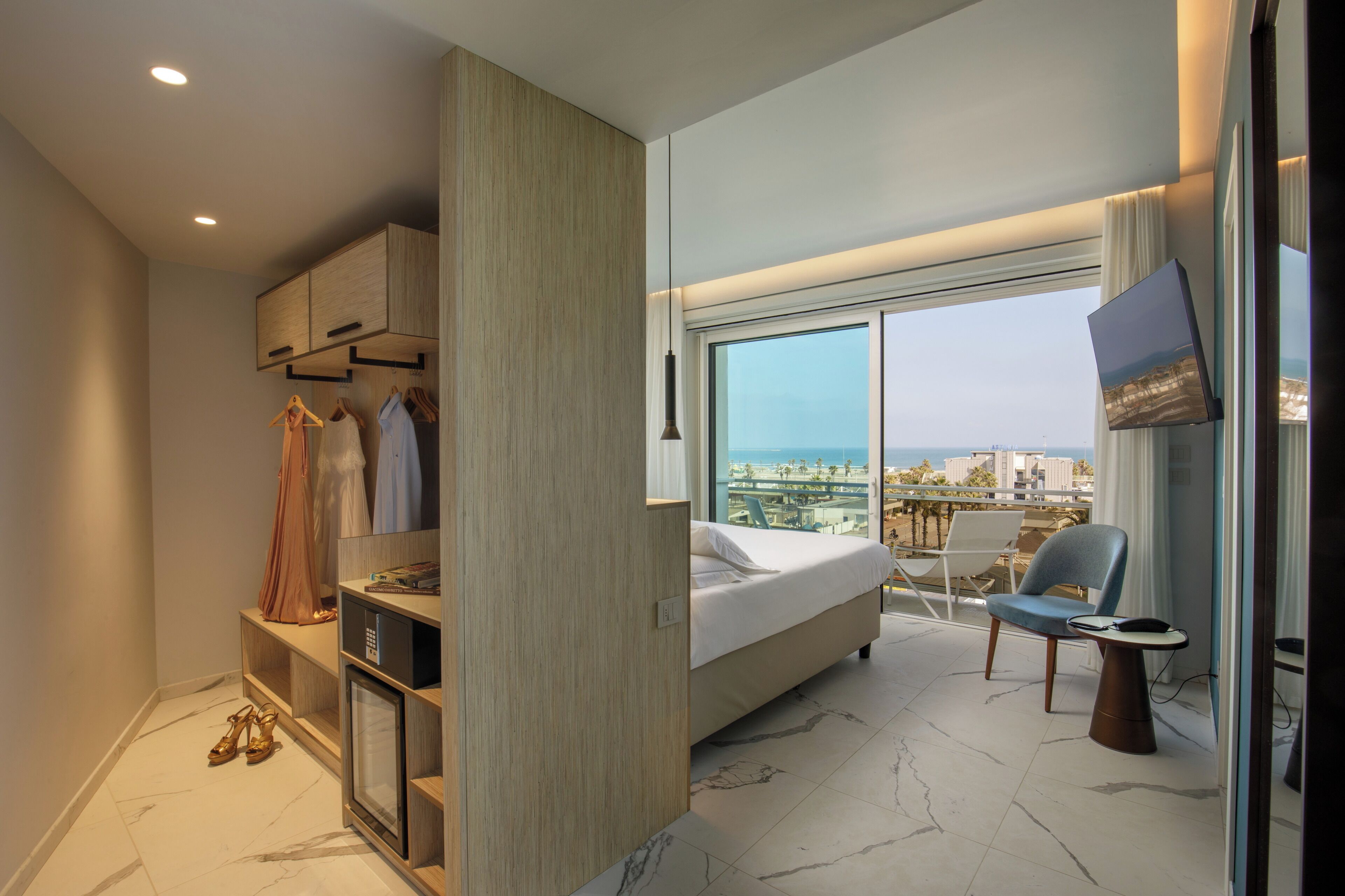 A modern hotel room with a bed, a seating area with a chair and table, a wardrobe with hanging clothes, and a large window offering a city view.