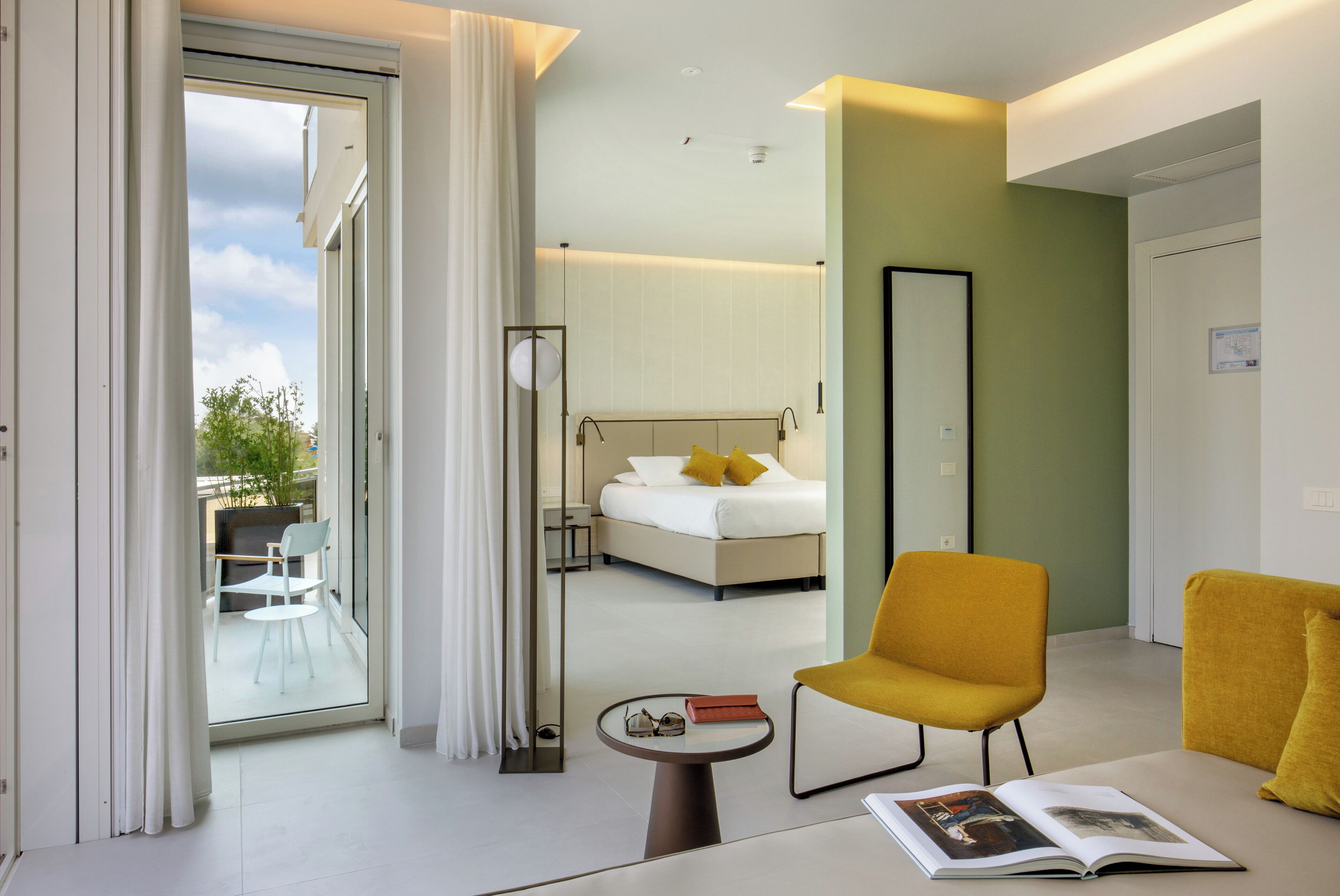 A modern hotel room with a bed, a yellow armchair, a small table with a magazine, and a large mirror.
