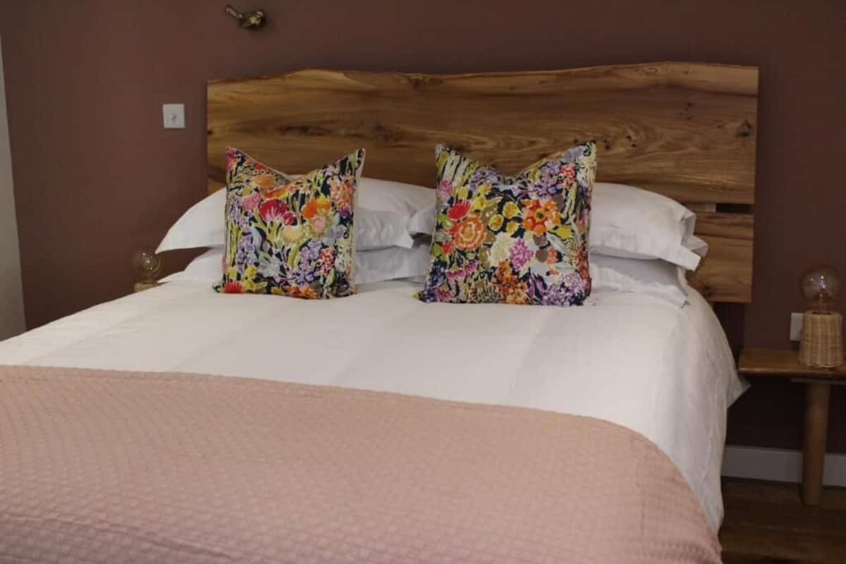 Una cama con cabecera de madera, dos almohadas estampadas con flores y una manta rosa.