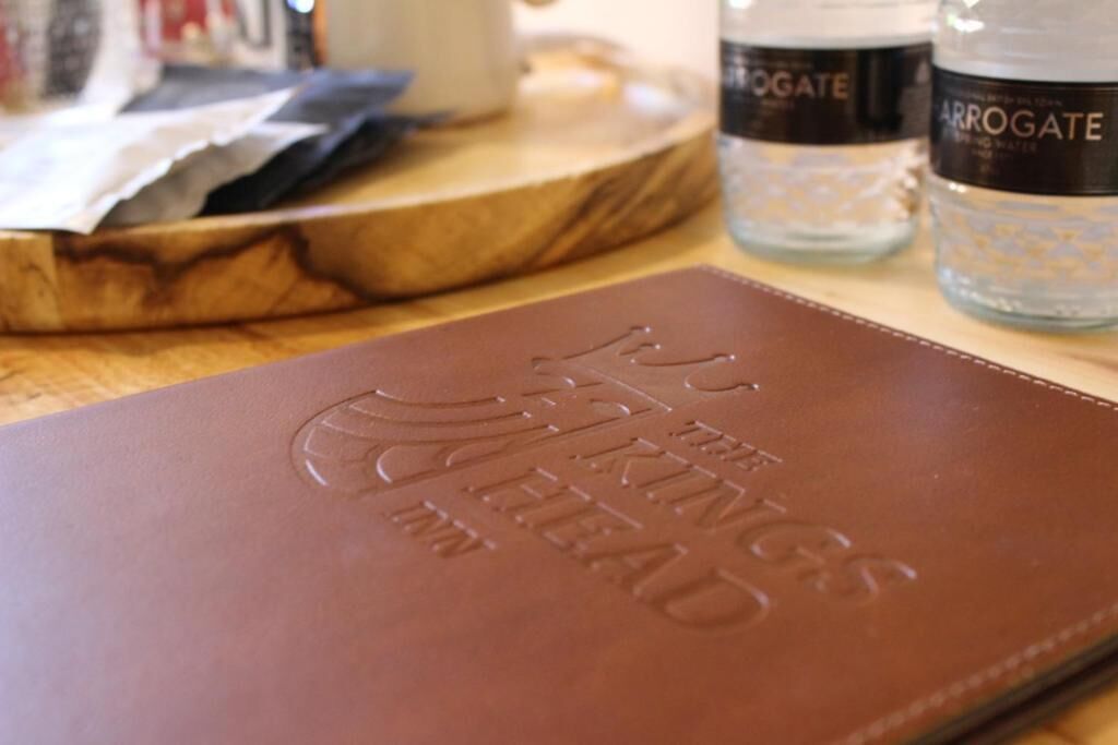 Un livre à la couverture en cuir marron, orné de caractères en relief, posé sur une surface en bois, avec deux bouteilles portant l’étiquette « ARROGATE » en arrière-plan.
