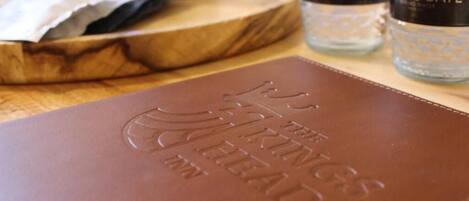 Un livre à la couverture en cuir marron, orné de caractères en relief, posé sur une surface en bois, avec deux bouteilles portant l’étiquette « ARROGATE » en arrière-plan.