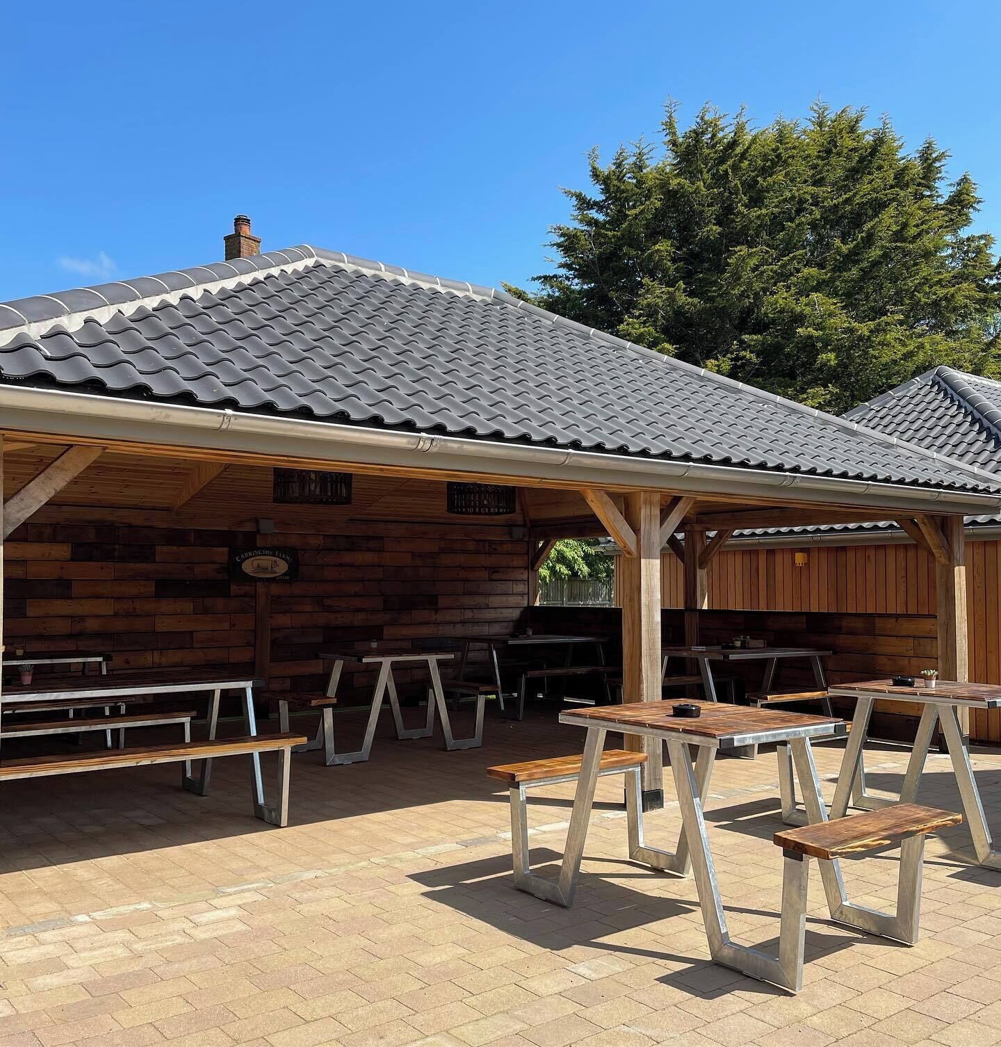 A covered outdoor seating area with wooden benches and tables, a tiled roof, and a wooden wall.