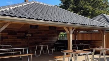 A covered outdoor seating area with wooden benches and tables, a tiled roof, and a wooden wall.