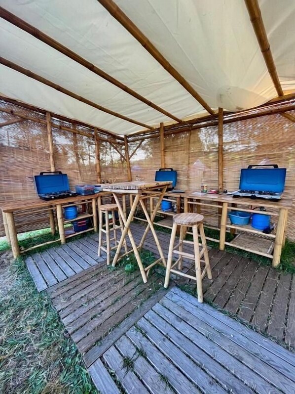 Een overdekt terras met houten tafels en stoelen, een centrale tafel met een blauwe koelbox, en een bamboe wand.