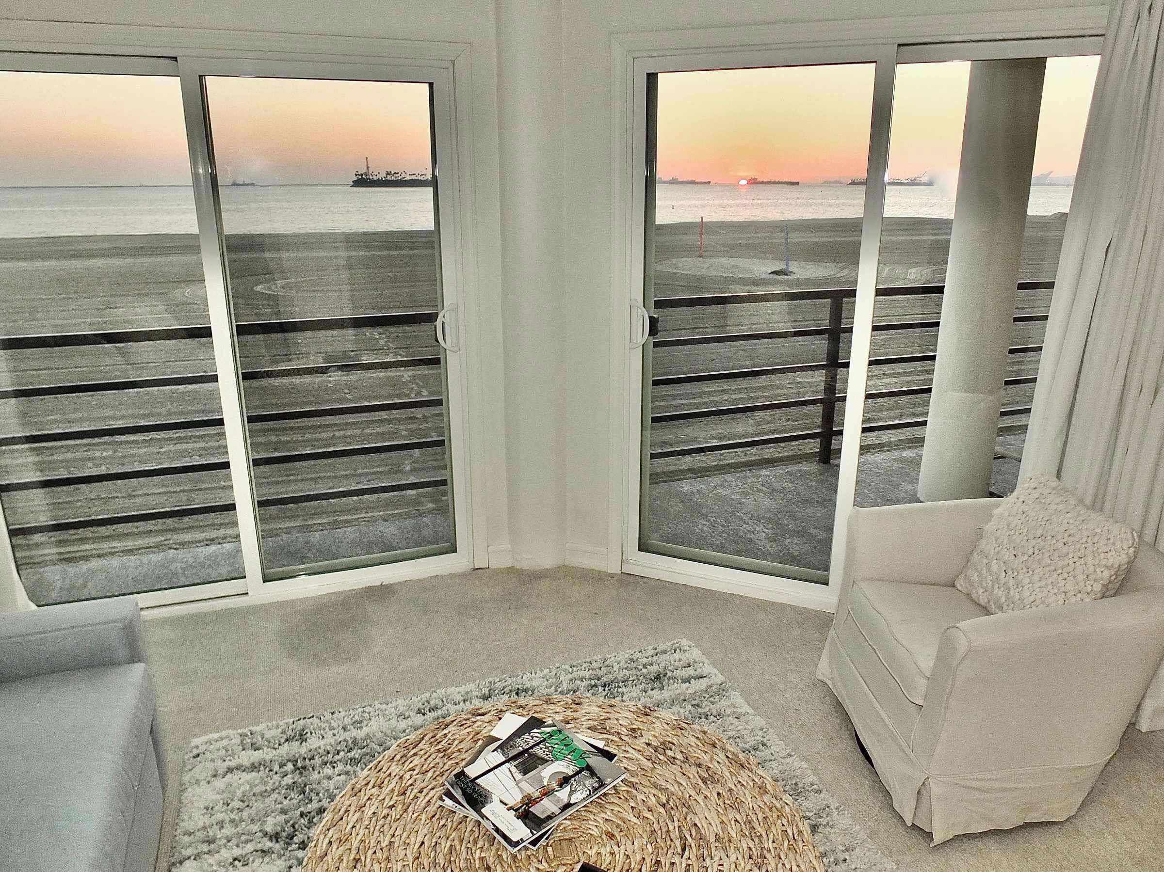 A room with a large sliding glass door leading to a balcony with a view of the sea and a ship. There is a round woven coffee table with magazines and a white armchair.
