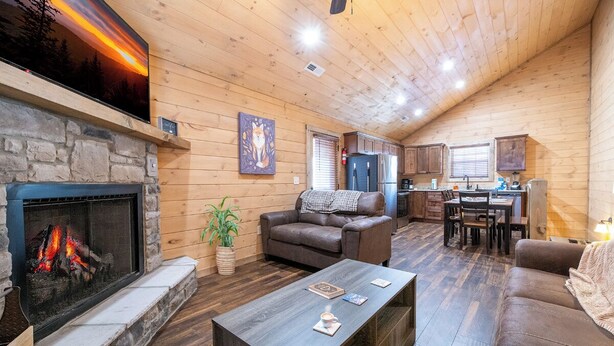A cozy cabin interior with a stone fireplace, wooden walls, a dining area, and a kitchen.