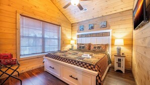 A cozy cabin-style bedroom with a wooden ceiling, a bed with a patterned bedspread, two bedside lamps, and a small cabinet.