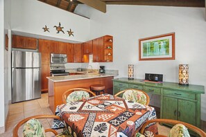 A kitchen with wooden cabinets, a refrigerator, a microwave, a coffee maker, and a dining area with a patterned tablecloth.