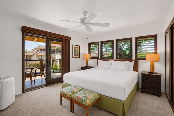 A hotel room with a bed, a ceiling fan, a window with blinds, a nightstand, a lamp, a chair, and a view of the outside.