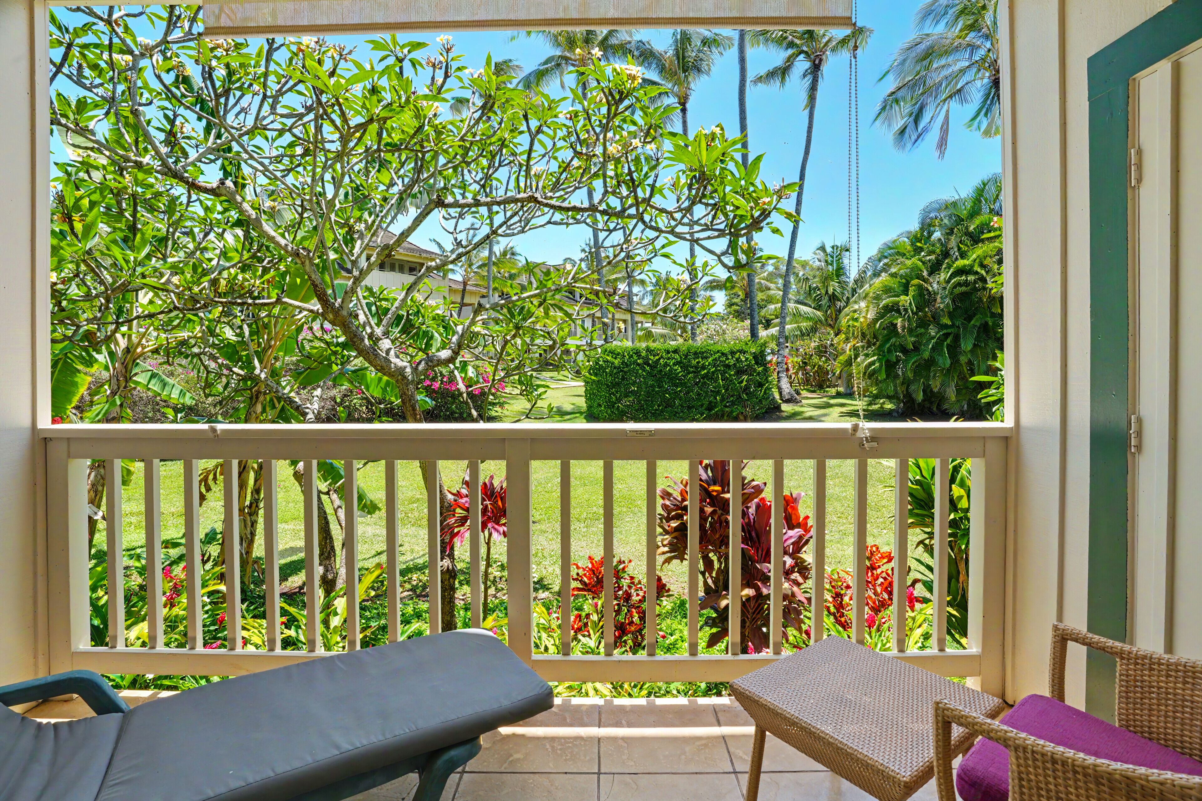 A balcony with wicker furniture, a view of a garden with various plants and a building in the background.