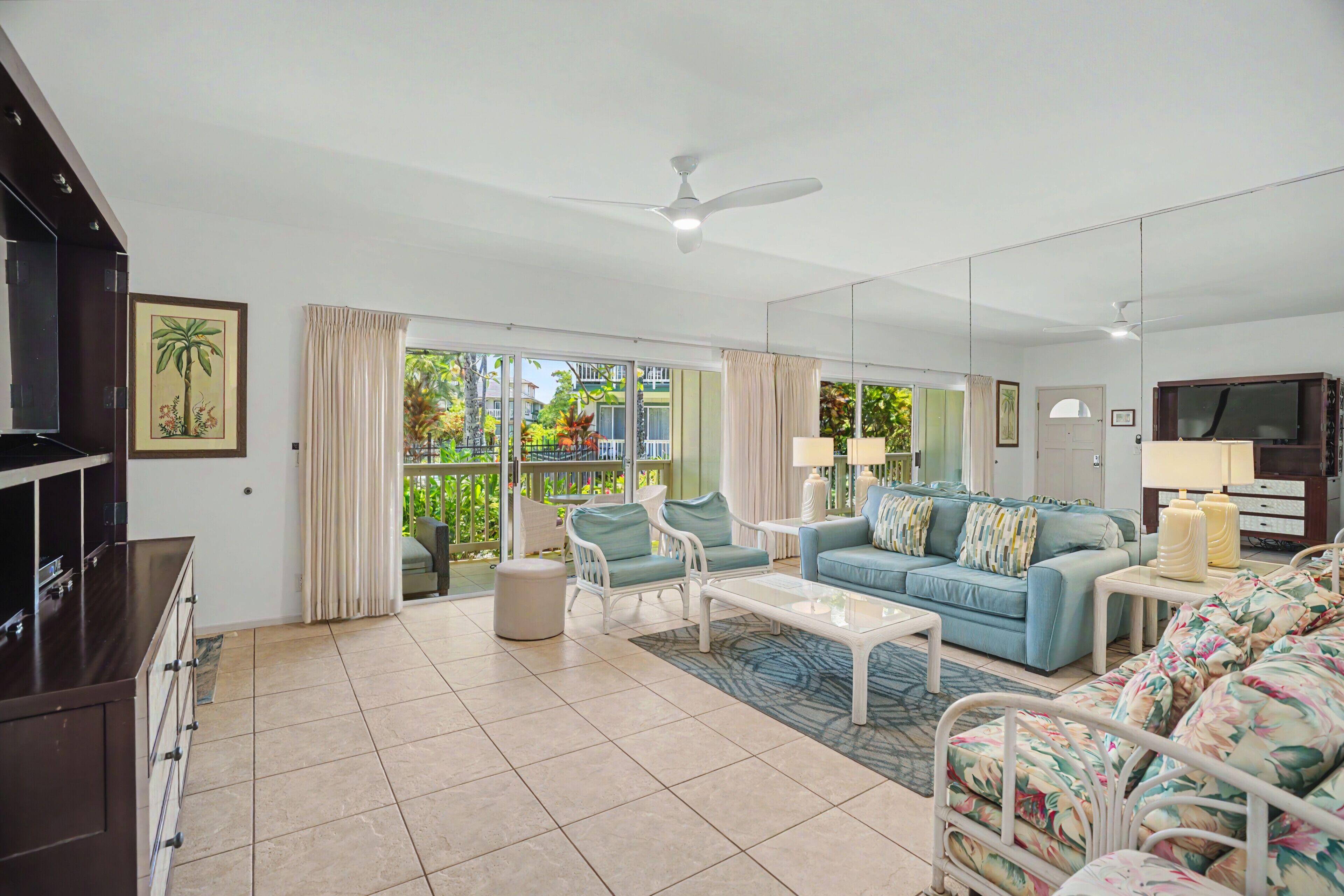 A spacious living area with a sofa, chairs, a coffee table, a TV, and a balcony with a view of greenery.