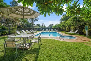 Zona de piscina con sillones de descanso y una mesa, rodeada de césped y árboles bien cuidados.