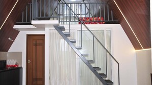 Un appartement moderne de style industriel, avec une porte en bois, un escalier doté de rampes en verre et un espace en mezzanine agrémenté d’un coussin rouge.