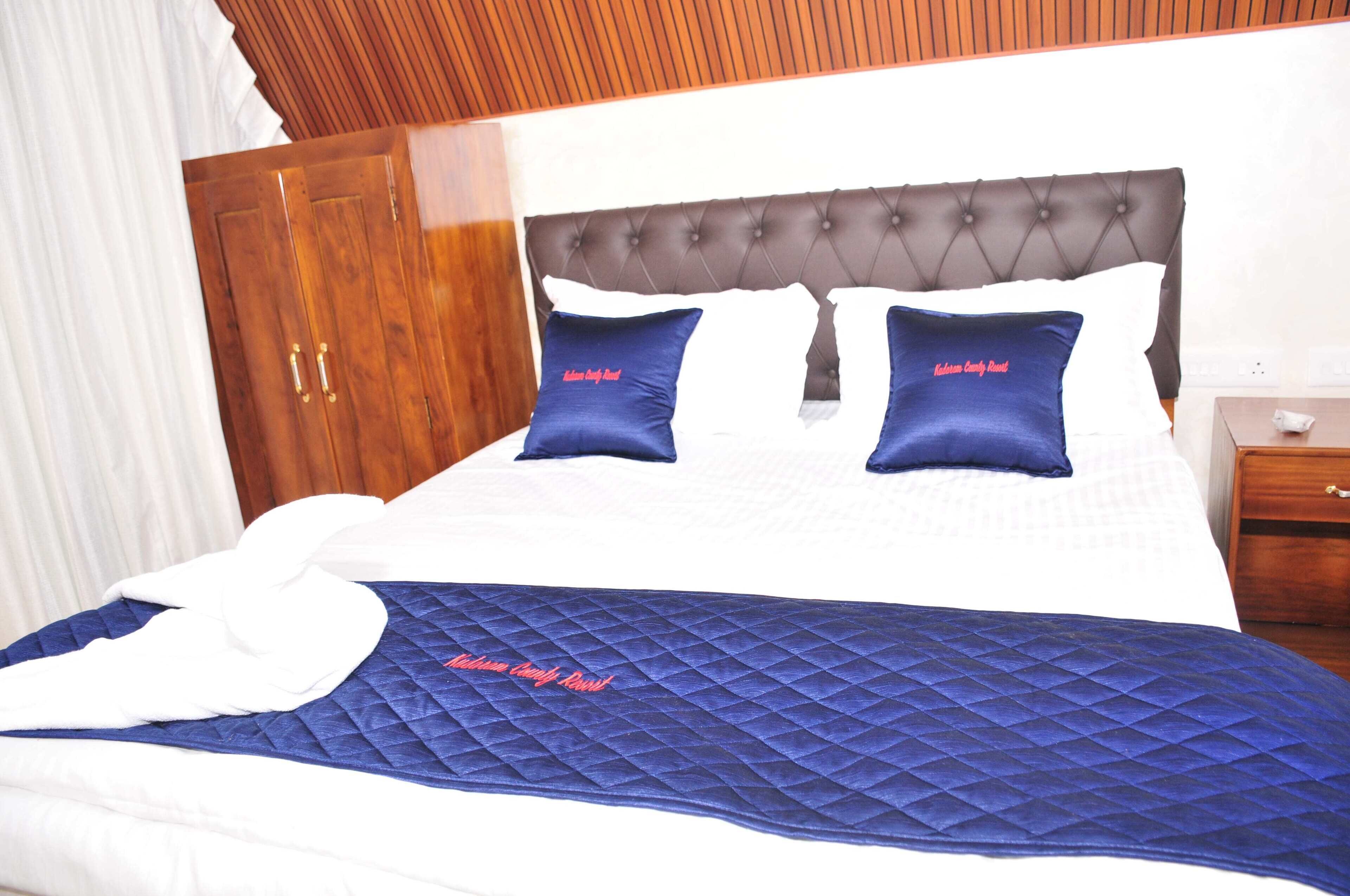 Una cama bien hecha con un edredón azul y dos almohadas, un armario de madera y una mesita de noche con una lámpara.
