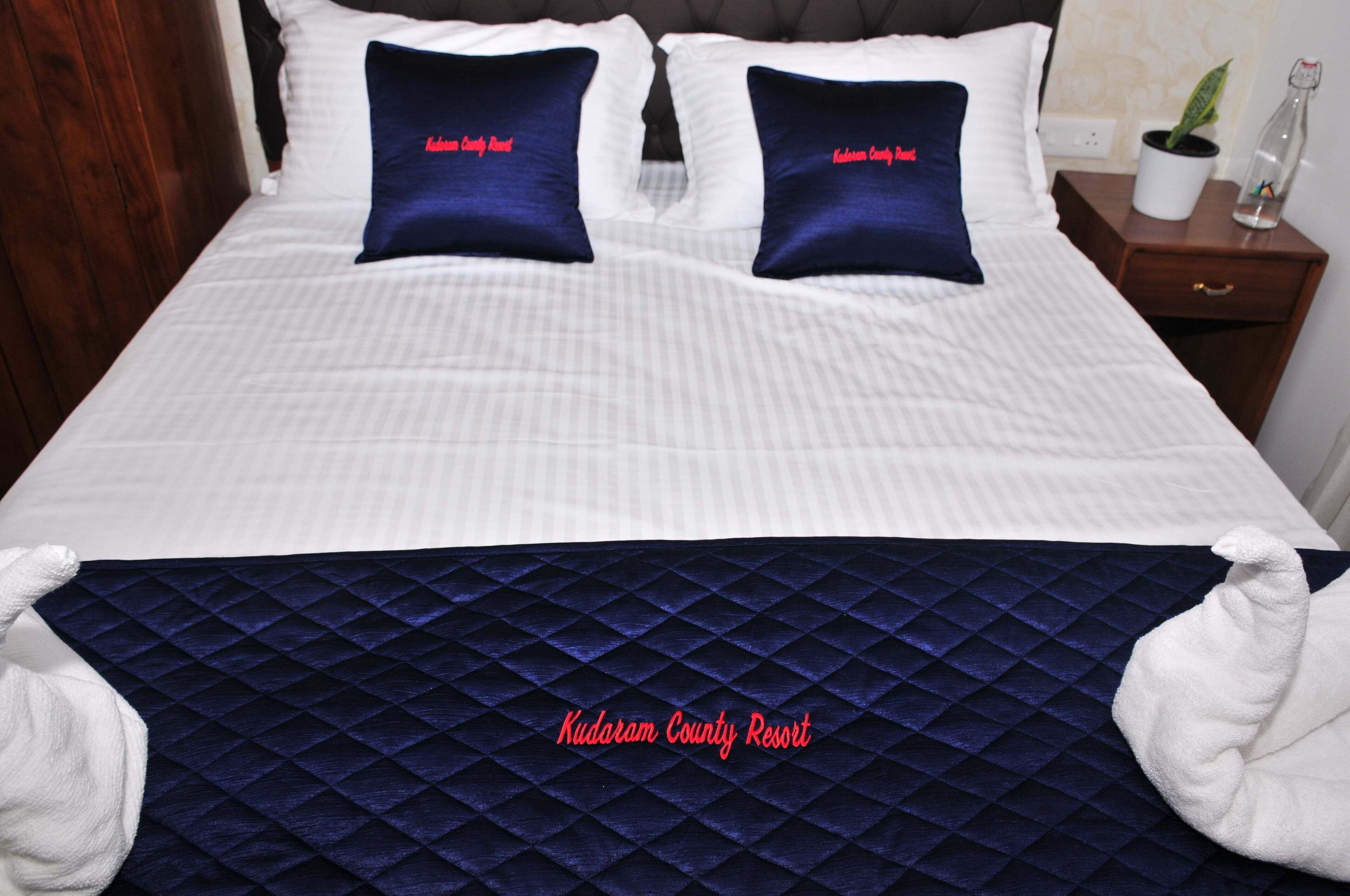 A neatly made bed with two pillows labeled "Kudoram County Resort," a dark blue quilted bedspread, and a bedside table with a plant and a bottle.