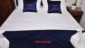A neatly made bed with two pillows labeled "Kudoram County Resort," a dark blue quilted bedspread, and a bedside table with a plant and a bottle.