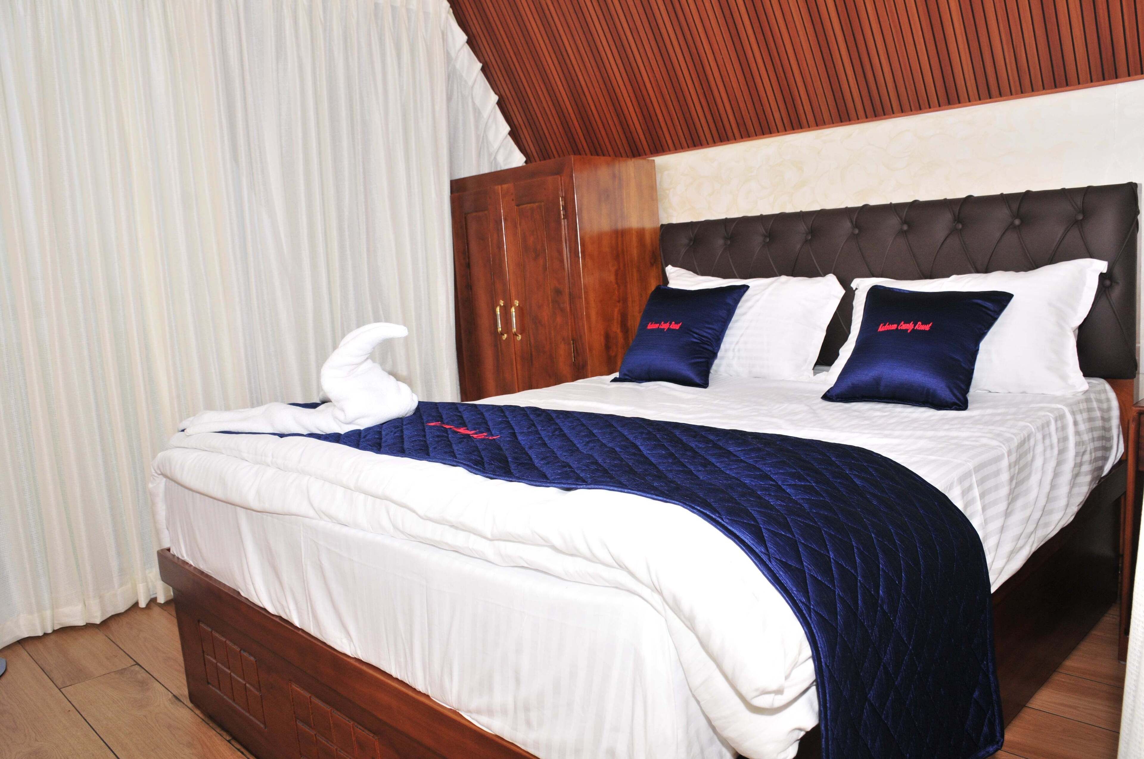 Una cama bien hecha con colcha azul y ropa de cama blanca, un armario de madera y un cabecero capitonado.