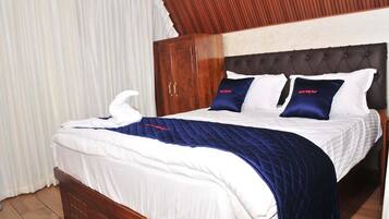 Una cama bien hecha con colcha azul y ropa de cama blanca, un armario de madera y un cabecero capitonado.
