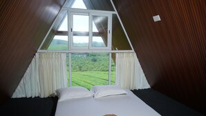 Une pièce triangulaire comprenant un lit, des rideaux blancs et une fenêtre donnant sur un paysage verdoyant.