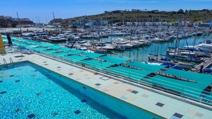 Un puerto deportivo con numerosas embarcaciones amarradas, una piscina y un paisaje montañoso al fondo.