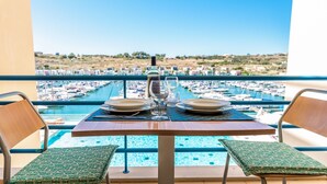 Una mesa de comedor con dos platos, una copa de vino y una botella, ubicada en un balcón con vistas a un puerto deportivo y una piscina.