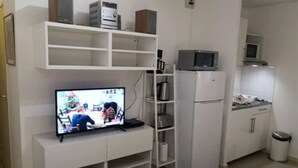 Un espacio habitable compacto con un televisor de pantalla plana sobre un mueble blanco, un microondas y una pequeña cocina con armarios y un cubo de basura.
