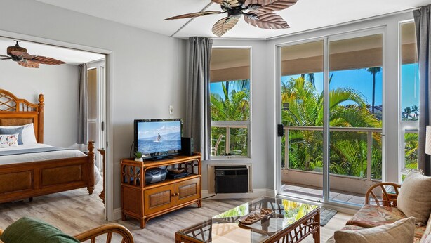 A spacious living room with a large TV, comfortable seating, a ceiling fan, and a balcony with tropical views.