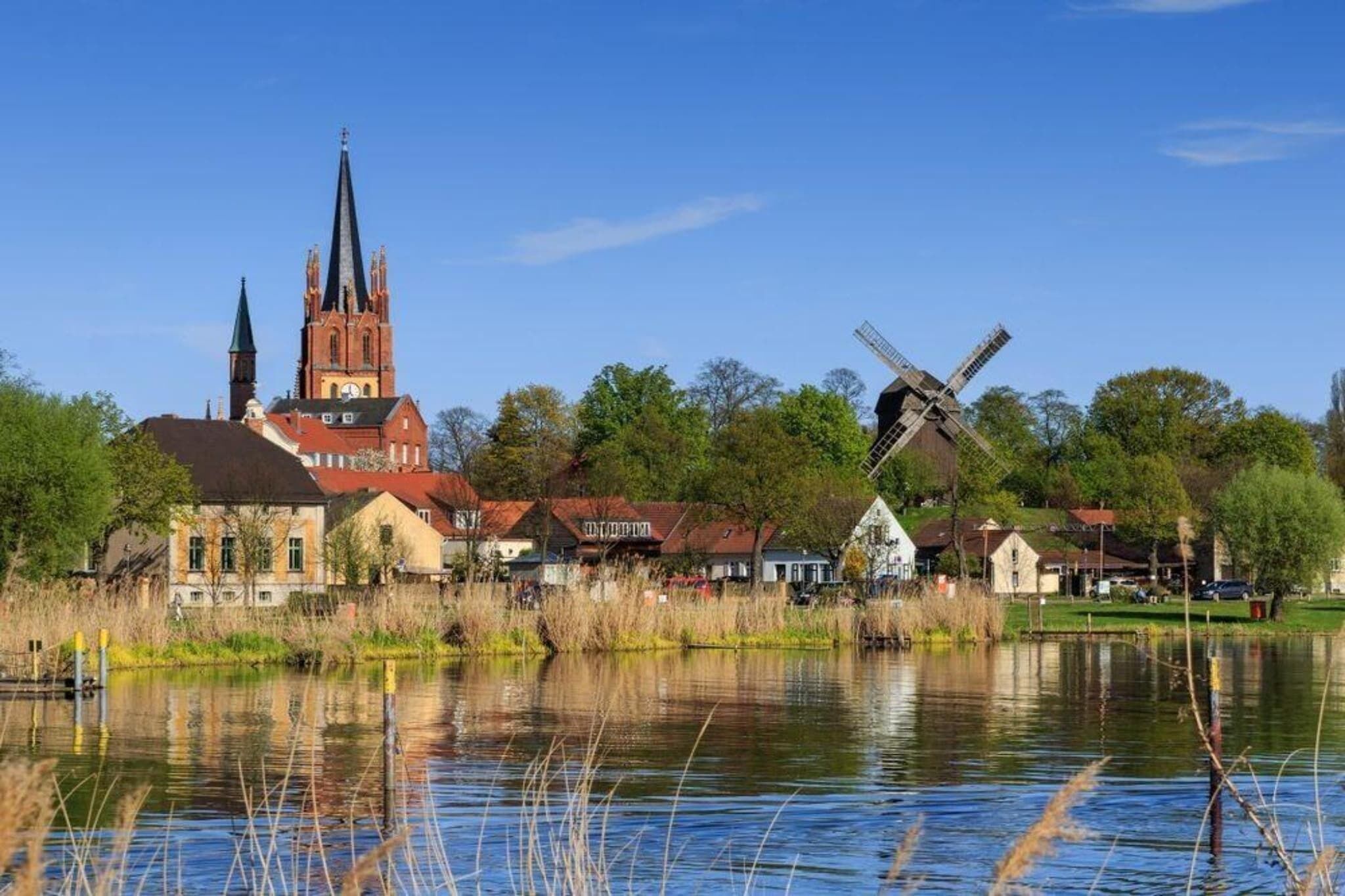 Une église dotée d’un flèche imposante, un moulin à vent traditionnel et une rivière paisible dans laquelle se reflètent les bâtiments.