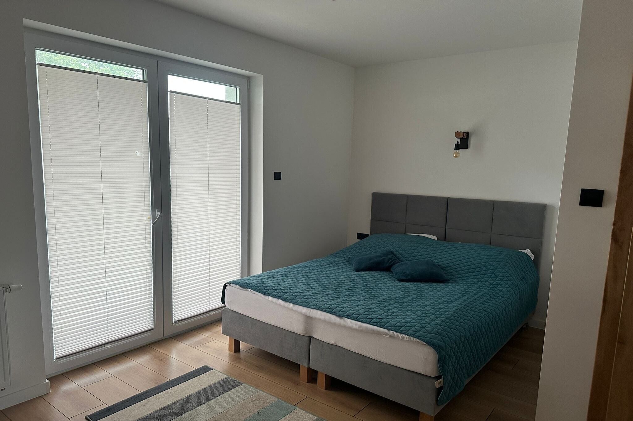 A bedroom with a bed, a grey headboard, a window with blinds, and a small wall-mounted light.