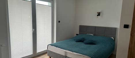 A bedroom with a bed, a grey headboard, a window with blinds, and a small wall-mounted light.