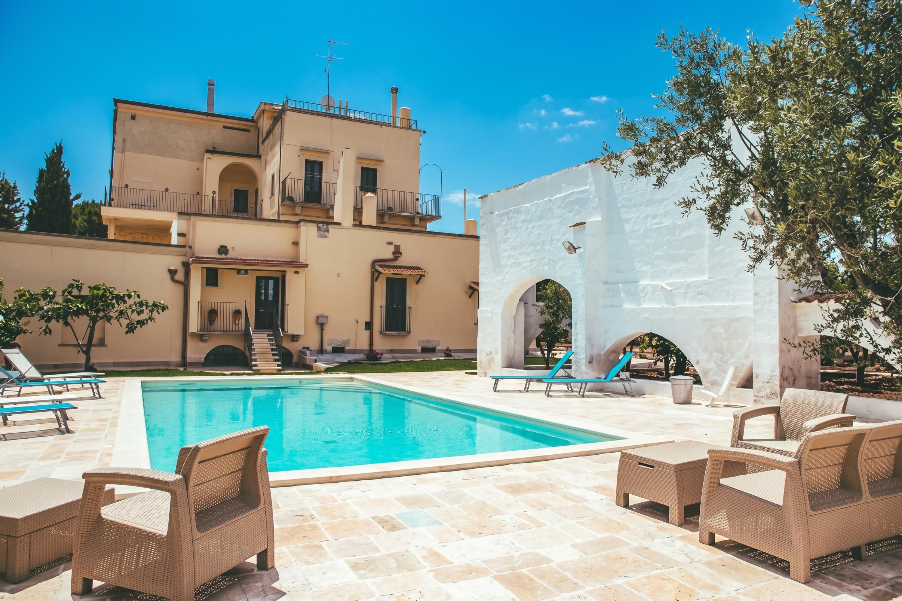 A swimming pool with lounge chairs, a white structure with arches, and a multi-story building in the background.