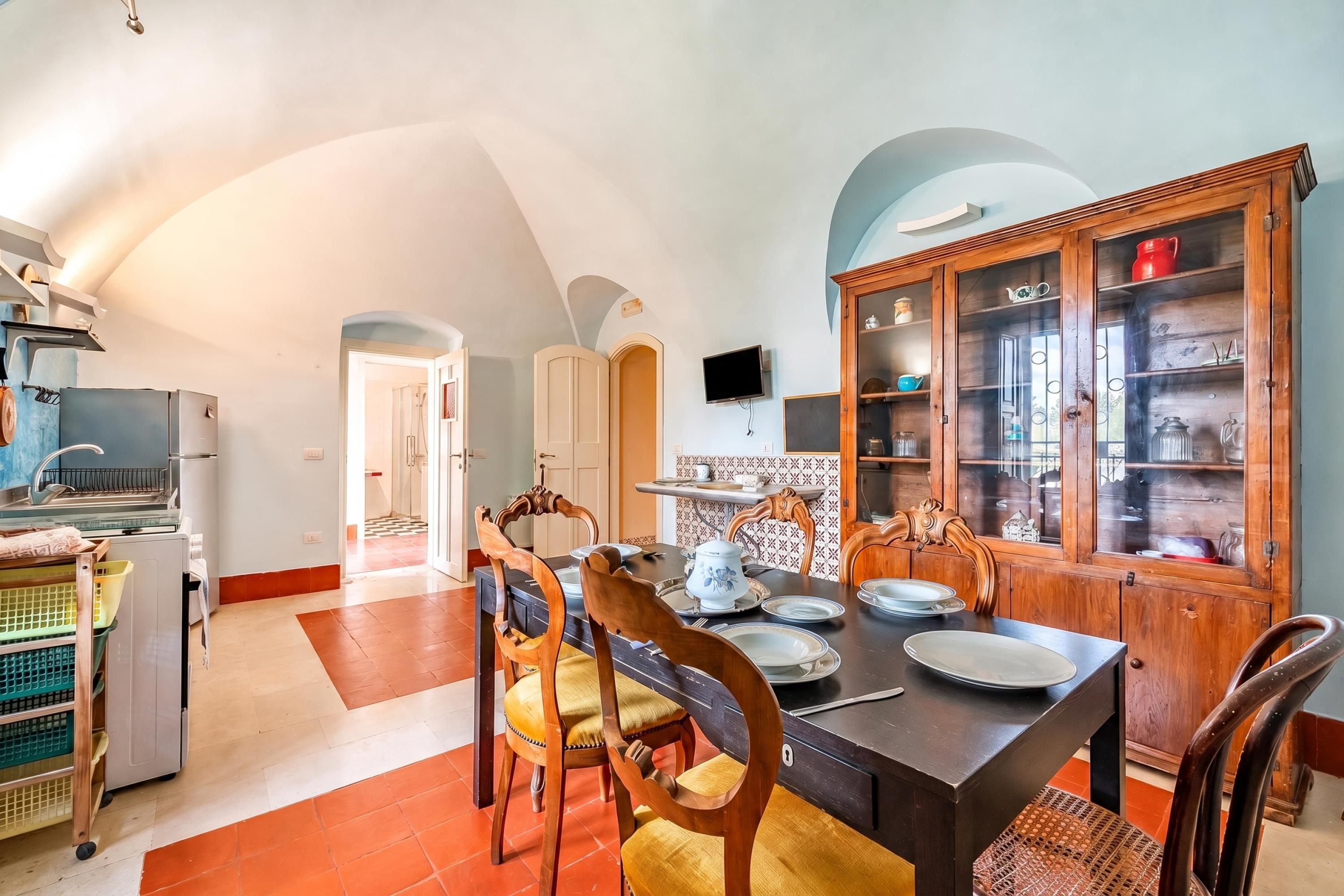 Cocina amplia con comedor, que cuenta con un mueble de madera grande con puertas de vidrio, horno empotrado y televisor montado en la pared.