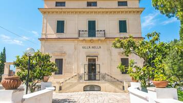 Um edifício com a placa 'VILLA NELLA' na entrada, um pátio com calçada de pedra e plantas em vasos ao redor.