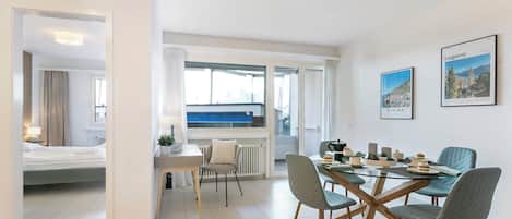 Et moderne hotelværelse med seng, spiseplads med bord og stole samt udsigt til blå himmel gennem store vinduer.